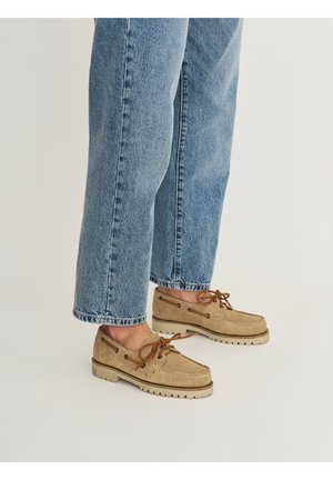 Persoon die lichtblauwe spijkerbroek en beige suède bootschoenen met bruine veters draagt, staand op een lichte achtergrond.