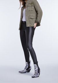 Pantalones de cuero negros, chaqueta verde oliva con dos bolsillos en el pecho y botas hasta el tobillo con un patrón de serpiente en blanco y negro.