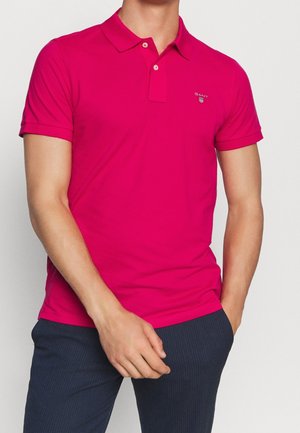 Man wearing a bright pink short-sleeve polo shirt with a small logo on the chest and dark pinstriped pants.