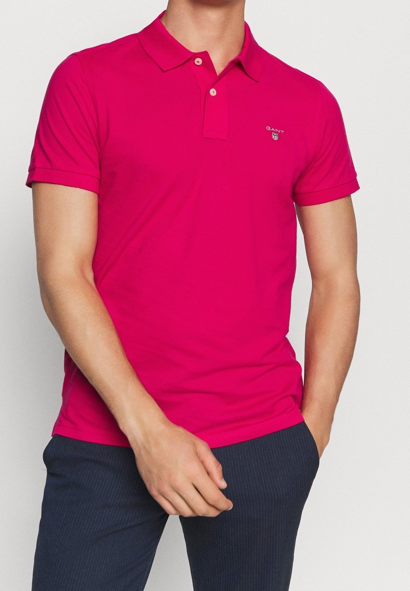 Homme portant un polo à manches courtes rose vif avec un petit logo sur la poitrine et un pantalon à fines rayures foncées.