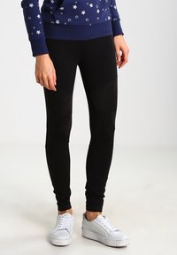 Svarta leggings med en åtsittande design som har en slät yta och knäpaneldetaljer, tillsammans med vita sneakers.