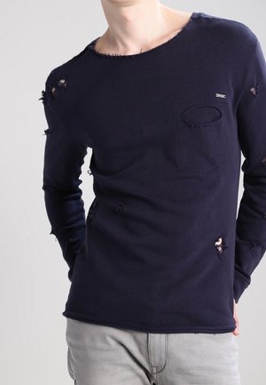 Mężczyzna w granatowej koszuli z długim rękawem z licznymi celowymi rozdarciami i jasnoszarych spodniach.