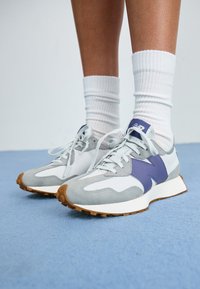 Scarpe sportive con upper in suede grigio e rete bianca, dettagli viola e suola in gomma. I lacci sono grigi, abbinati a calze bianche a coste.