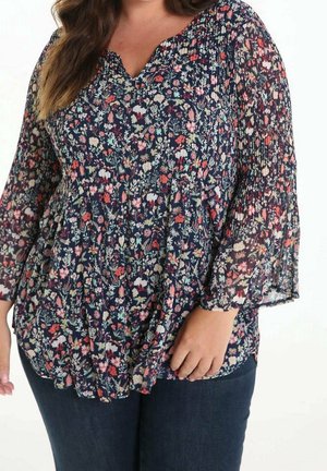 Vrouw draagt een donkerblauw gebloemd blouse met geplooide taille en halfdoorzichtige lange mouwen, gecombineerd met donkerblauwe jeans.