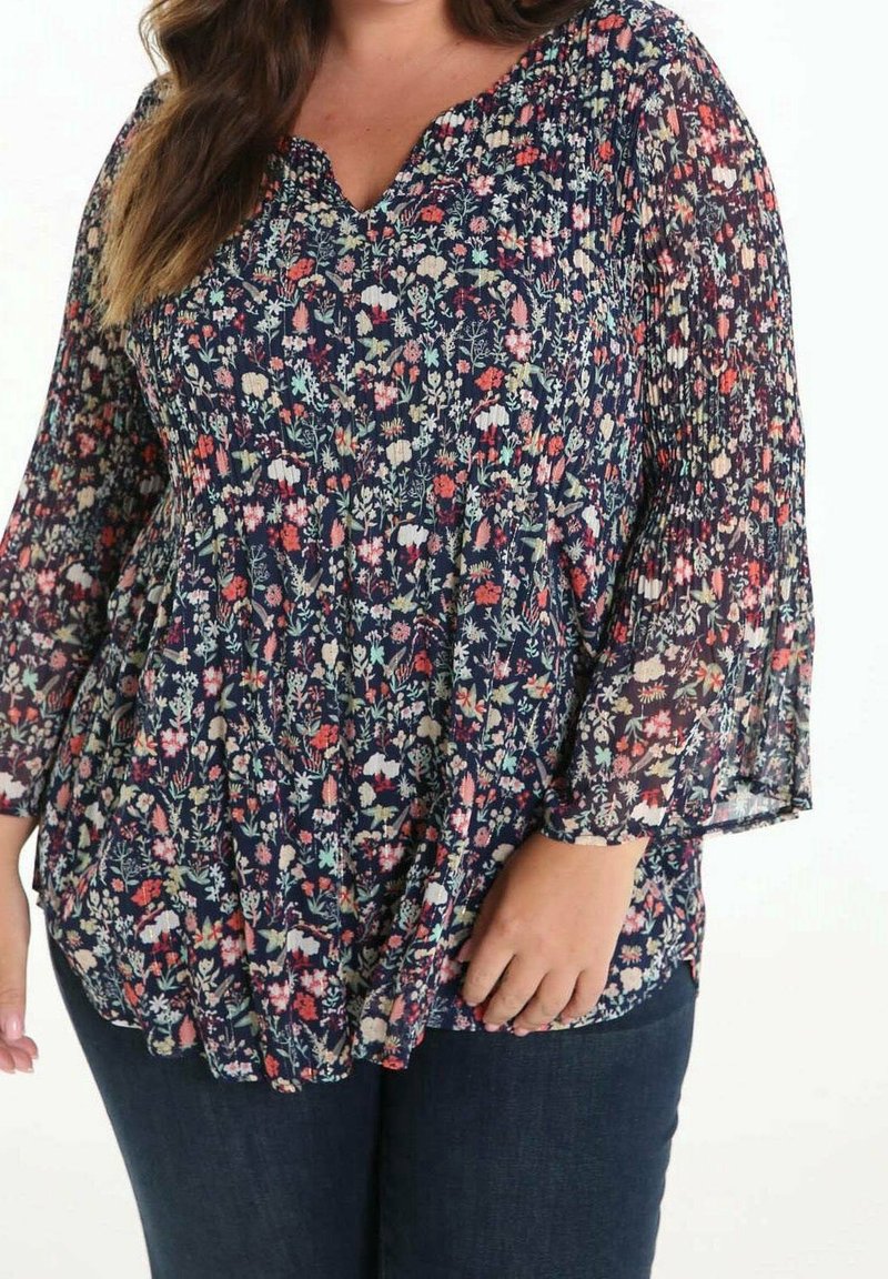 Femme portant un chemisier bleu foncé à motifs floraux avec taille froncée et manches longues semi-transparentes, associé à un jean bleu foncé.