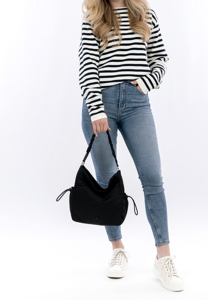 Zwarte stoffen hobo tas met gevlochten handvat en trekkoordaccenten. Model draagt een gestreepte lange mouw top en blauwe skinny jeans.