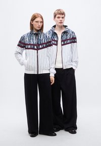 Δύο άτομα φορούν ίδια ζακέτα με φερμουάρ, διακοσμημένη με μπλε και λευκό μοτίβο και μπορντό ρίγες. Συνδυάζουν τις ζακέτες με μαύρο παντελόνι φαρδύ.