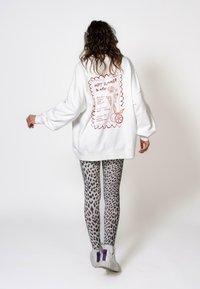Felpa bianca oversize con grafica rossa sul retro, abbinata a leggings grigi con stampa leopardata e sneakers bianche con accenti viola.