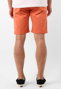 Shorts en coton orange avec un ourlet roulé, portés au-dessus du genou. Chaussures noires avec des semelles blanches ancrent la tenue. Fond blanc uni.