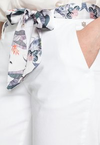 White trousers with a smooth texture, featuring a patterned fabric belt tied at the waist. The belt displays floral and leaf designs in pastel colors.