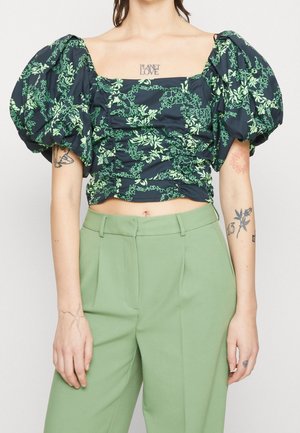 Kvinde iført en sort crop top med grøn blomstermønster og puffede ærmer, kombineret med højttaljede, lysegrønne bukser.