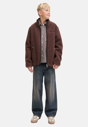 Veste en polaire marron avec un col montant, des poches zippées et une coupe décontractée, portée sur une chemise à carreaux et assortie à un jeans bleu ample.