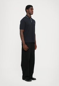 Marine polo skjorte med krage og to-knaps placket, med hvite detaljer. Kombinert med løst sittende svarte bukser og svarte sko.
