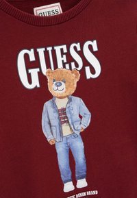 Sweat-shirt bordeaux arborant un motif d'un ours portant une veste en jean et une chemise beige, avec le mot "GUESS" affiché en évidence au-dessus.