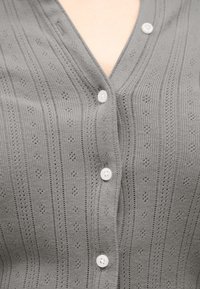 Cardigan en maille gris avec de petits trous décoratifs et des boutons blancs fermés sur une peau nue.