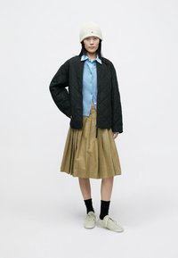 Schwarze gesteppte Jacke über einem hellblauen Hemd mit Knöpfen, kombiniert mit einem plissierten khakifarbenen Rock und cremefarbenen Turnschuhen, dazu eine weiße Mütze.