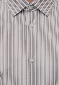 Gros plan d'une chemise blanche avec des rayures verticales marron et grises, montrant le col et trois boutons blancs.
