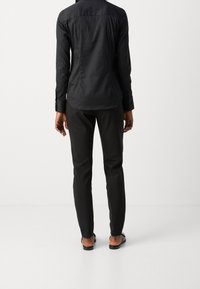 Camicia nera con bottoni, maniche lunghe, abbinata a pantaloni neri. Il materiale sembra liscio e aderente, con un design minimalista.