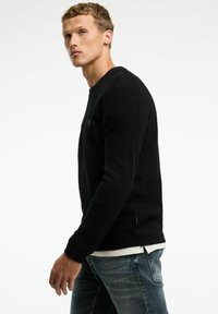 Schwarzer strukturierter Strickpullover mit gerippten Ärmeln, Rundhalsausschnitt und dezentem Logo-Detail. Über einem hellen Hemd getragen und mit blauen Jeans kombiniert.