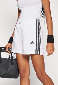 Beli šorti z črnimi stranskimi črtami, elastičnim pasom in Adidasovim logotipom. V kombinaciji s črnimi zapestnicami in sivo torbo.