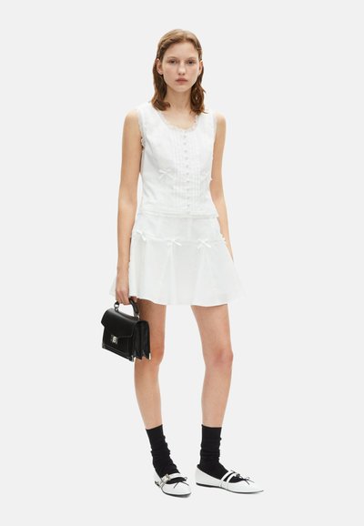 Robe blanche sans manches avec bord en dentelle, détails de boutons et accents de nœud ; portée avec un sac à main noir, des chaussettes jusqu'aux genoux et des chaussures plates.