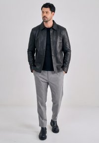 Mann mit dunklem Haar und Bart, der eine schwarze Lederjacke, ein schwarzes Hemd, graue Hosen und schwarze Stiefel trägt und vor einem einfachen Hintergrund steht.