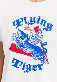 Hvid t-shirt med et grafisk design af en blå løve med vinger, fed blå tekst "Flying Tiger" og en rød geometrisk accent.