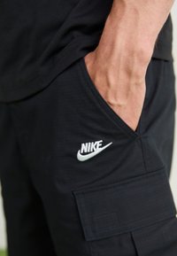 Černé cargo kalhoty s boční kapsou, texturovanou tkaninou a bílým vyšitým logem Nike na přední straně.