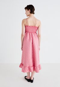 Pink polka dot midi dress with a smocked bodice, thin straps, and ruffled hem. Smooth fabric with a shiny finish. Black slip-on shoes worn.