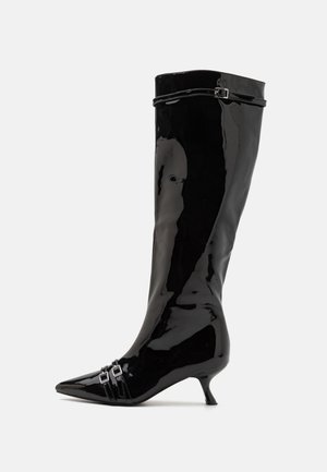 Botă neagră din piele lacuită, până la genunchi, cu vârf ascuțit, toc mic și accente cu două catarame aproape de gleznă. Finisaj neted și lucios.