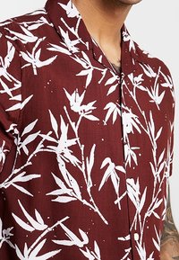 Camicia a maniche corte color bordeaux con motivo di foglie di bambù bianche, colletto e bottoni, indossata da una persona con il braccio e il collo tatuati visibili.