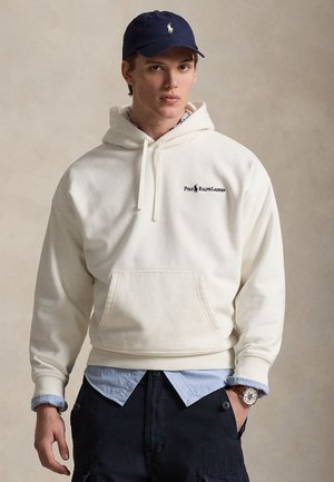 Kreemjas kapuutsiga dressipluus esitasku ja paeladega, millel on must logo. Kandmisel hele sinise särgi kohal; kombineeritud tumedate pükste ja käekellaga.