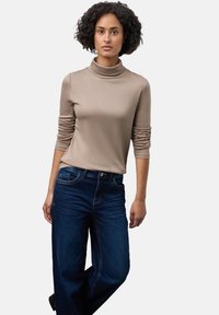 Beige Rollkragenoberteil mit langen Ärmeln, das eine glatte Textur aufweist. Kombiniert mit dunkelblauen weit geschnittenen Jeans mit Steppnähten.