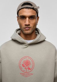 Beige hoodie met een grote rode rozenafbeelding en tekst aan de voorkant. Voorzien van een trekkoord capuchon en een casual pasvorm. Katoenen materiaal.