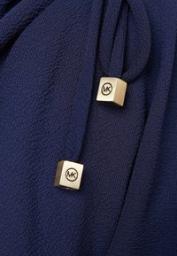 Tissu texturé bleu marine avec deux extrémités carrées en métal doré sur les cordons, chacune gravée du logo "MK".