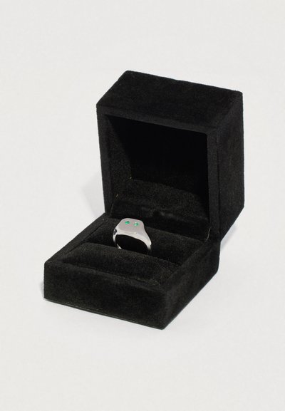 Fekete bársony nyitott gyűrűtartó doboz, amelyben egy ezüst gyűrű van két kis zöld drágakővel a tetején, egyszerű világos háttér előtt.