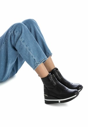 Stivali ankle a zeppa neri con chiusura a zip, tomaia in pelle liscia, punta rotonda e una striscia bianca sul fondo. Pantaloni in denim parzialmente visibili.
