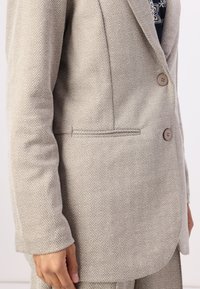 Beige herringbone musterniga bleiser, mis on valmistatud tekstuursetest kangast, kahe nupuga ja ühe rinnataskuga, kohandatud lõikega ning sileda voodriga.