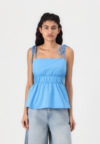 Blue peplum top with adjustable tie shoulder straps, gathered waist, and smooth texture. Paired with light denim wide-leg jeans.