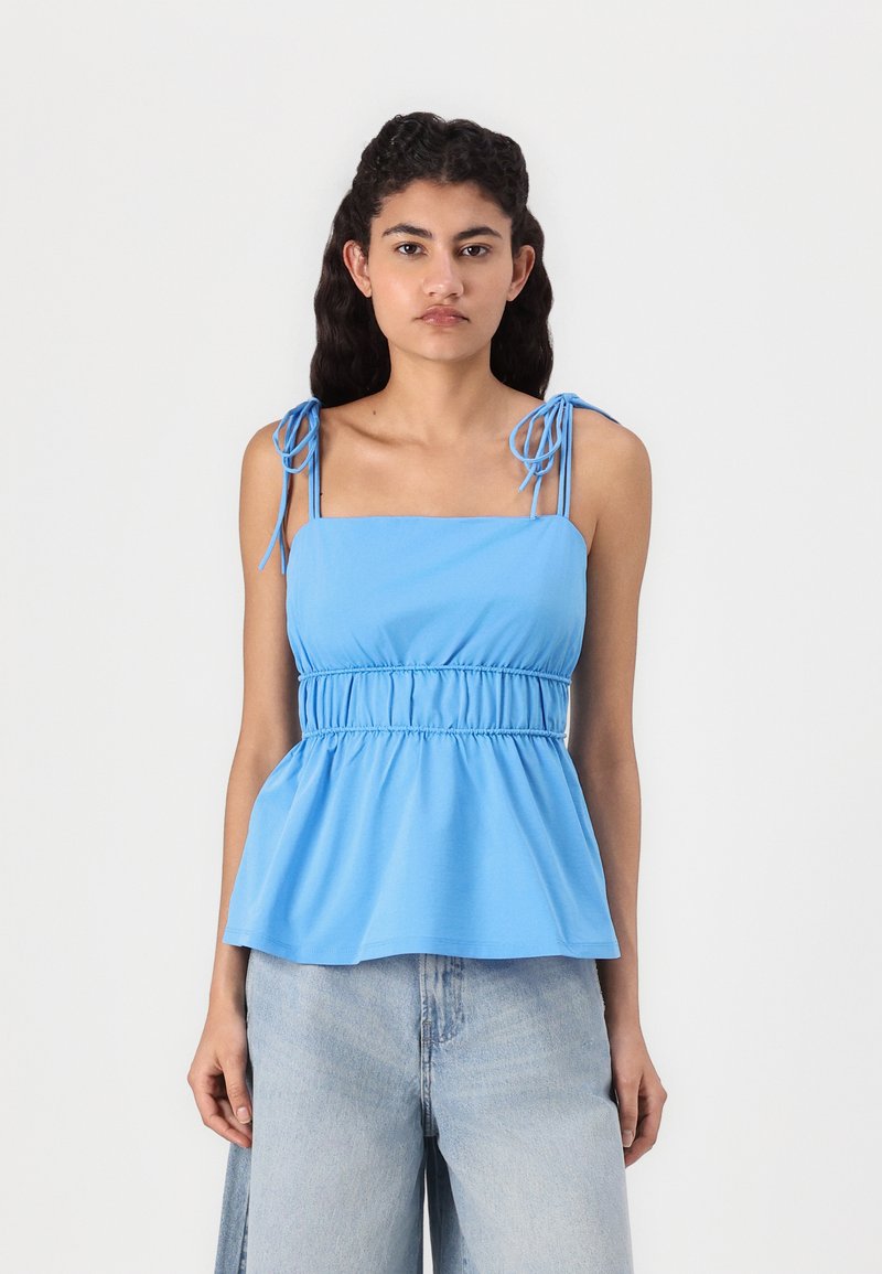 Blue peplum top with adjustable tie shoulder straps, gathered waist, and smooth texture. Paired with light denim wide-leg jeans.