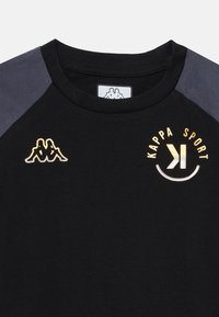Juodas trumpomis rankovėmis marškinėlis su tamsiai pilkais raglan rankovėmis. Priekyje atspausdinta auksinė logotipas ir tekstas "KAPPA SPORT". Lygi medvilnės medžiaga.