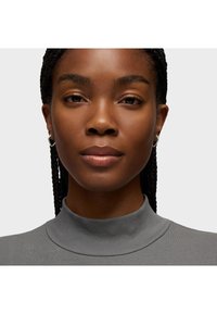 Mujer joven con el cabello trenzado, usando aretes de aro plateados y una camiseta gris de cuello alto, mirando con calma hacia adelante contra un fondo liso.