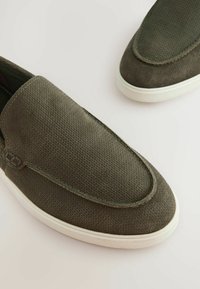 Maslinasto zelene slip-on cipele od teksturiranog materijala, s okruglim vrhom i belim gumiranim potplatima, koje imaju suptilan dijamantni uzorak i ušivene naglaske.