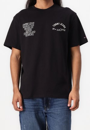 Mężczyzna w czarnej koszulce Tommy Jeans z białym nadrukiem wymieniającym dzielnice Nowego Jorku oraz szczegóły marki, w połączeniu z niebieskimi dżinsami.