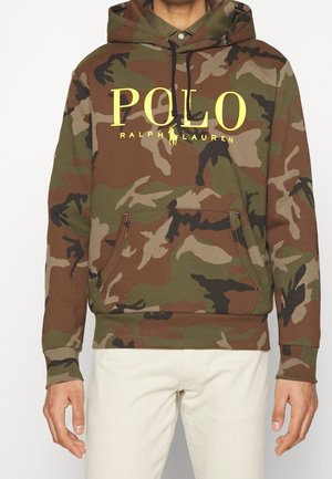 Mężczyzna noszący zieloną i brązową bluzę z kapturem w kamuflaż z żółtym napisem „POLO Ralph Lauren” i logo, w połączeniu z jasnobeżowymi spodniami.