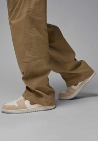 Lys brune baggy bukser med sidelommer og et lille broderet logo, parret med beige sneakers med lysebrune detaljer og en tekstureret sål.