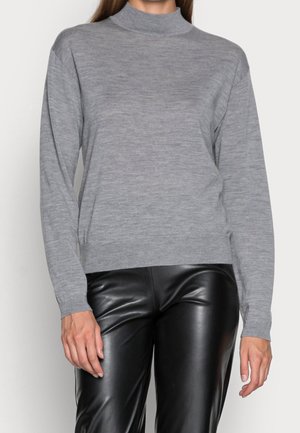 Femme portant un pull à col roulé gris clair à manches longues et un pantalon en cuir noir, debout devant un fond uni.