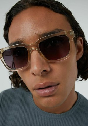 Transparante, vierkante zonnebril met donkere lenzen. Het montuur is lichtbeige met subtiele hardware-details. Stevige textuur tegen een neutrale achtergrond.