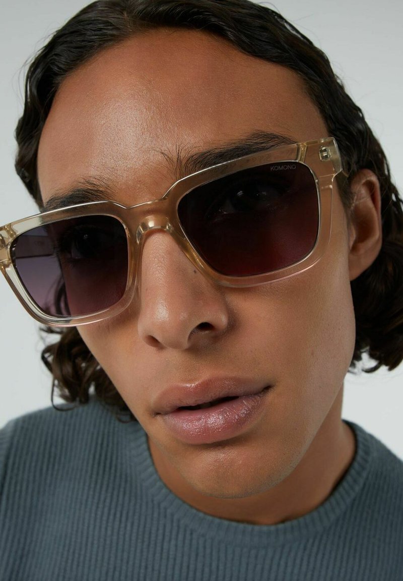 Transparante, vierkante zonnebril met donkere lenzen. Het montuur is lichtbeige met subtiele hardware-details. Stevige textuur tegen een neutrale achtergrond.