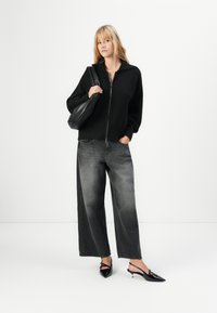 Ung kvinne iført en svart glidelåsgenser, vide falmede svarte jeans, svarte slingback-hæler, og en svart lær skulderveske.